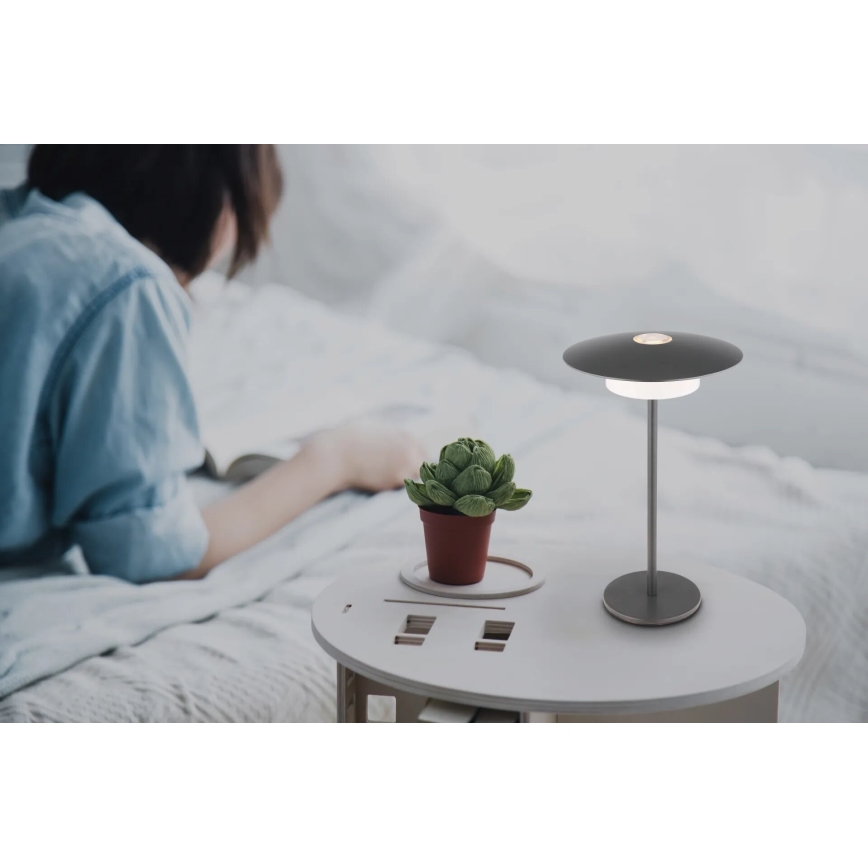 Stolová lampa s diskovým tienidlom svieti na nočnom stolíku. Žena leží v posteli a číta knihu. Na stolíku je tiež malý kvetináč so sukulentom.