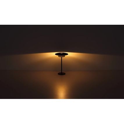 Stojacia lampa svieti. Má čierny kovový stojan a plochý tienidlo. Lampa stojí na svetlej podlahe pred tmavou stenou.