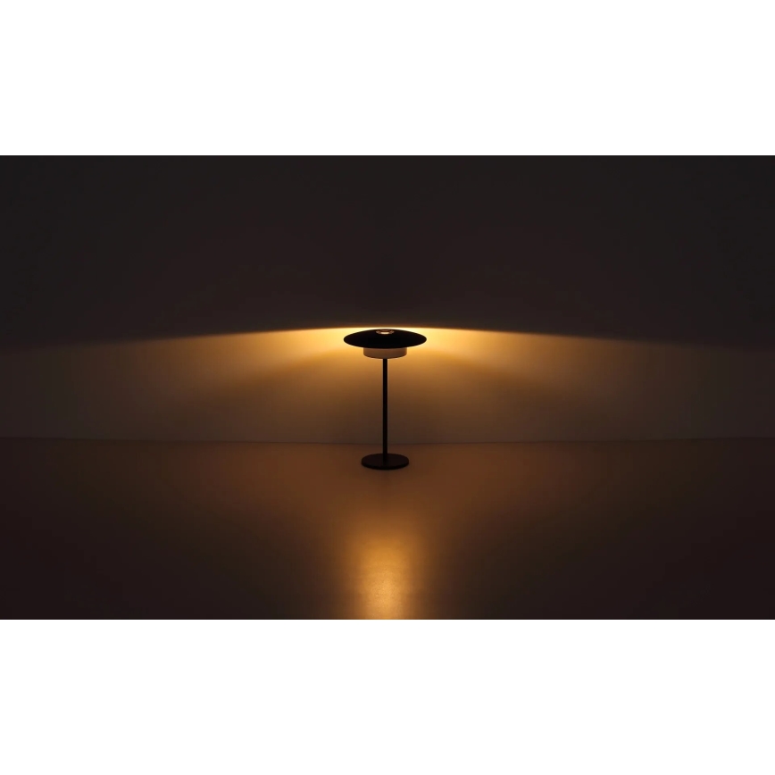 Stojacia lampa svieti. Má čierny kovový stojan a plochý tienidlo. Lampa stojí na svetlej podlahe pred tmavou stenou.