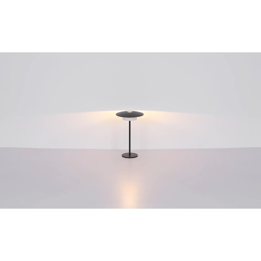 Stolová lampa s čiernym tienidlom v tvare taniera svieti na svetlej ploche. Lampa je umiestnená na svetlej podlahe pred svetlou stenou.