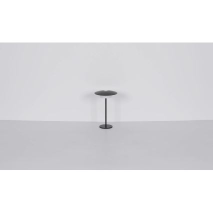 Stolová lampa s tmavosivým tienidlom v tvare disku stojí na tenkej čiernej nohe s kruhovou základňou. Lampa je umiestnená na svetlej podlahe pred bielou stenou.