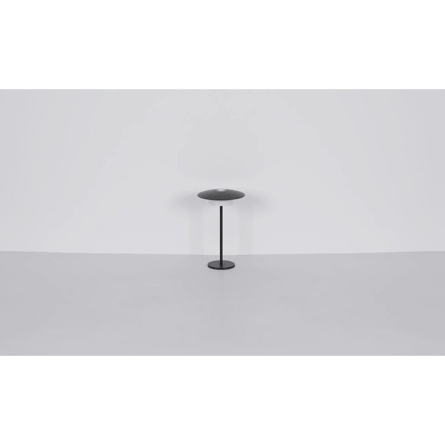 Stolová lampa s tmavosivým tienidlom v tvare disku stojí na tenkej čiernej nohe s kruhovou základňou. Lampa je umiestnená na svetlej podlahe pred bielou stenou.
