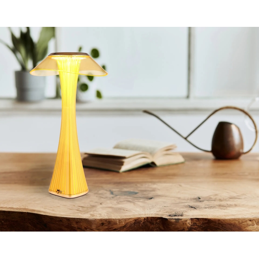 Stolová lampa s tvarom huby svieti na drevenom stole. Vedľa nej leží kniha a záhradná konvička.