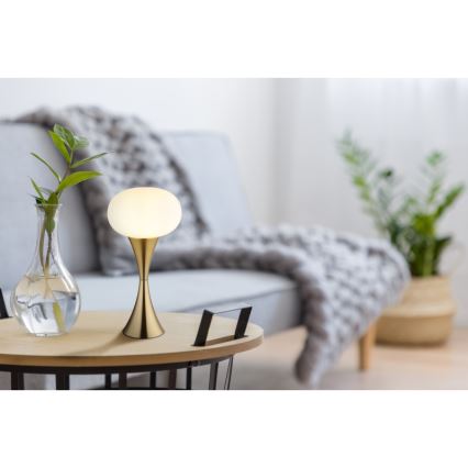 Stolná lampa s mliečnobielym tienidlom v tvare gule a zlatým podstavcom svieti na drevenom odkladacím stolíku. Lampa sa nachádza v obývacej izbe s pohovkou a rastlinami.