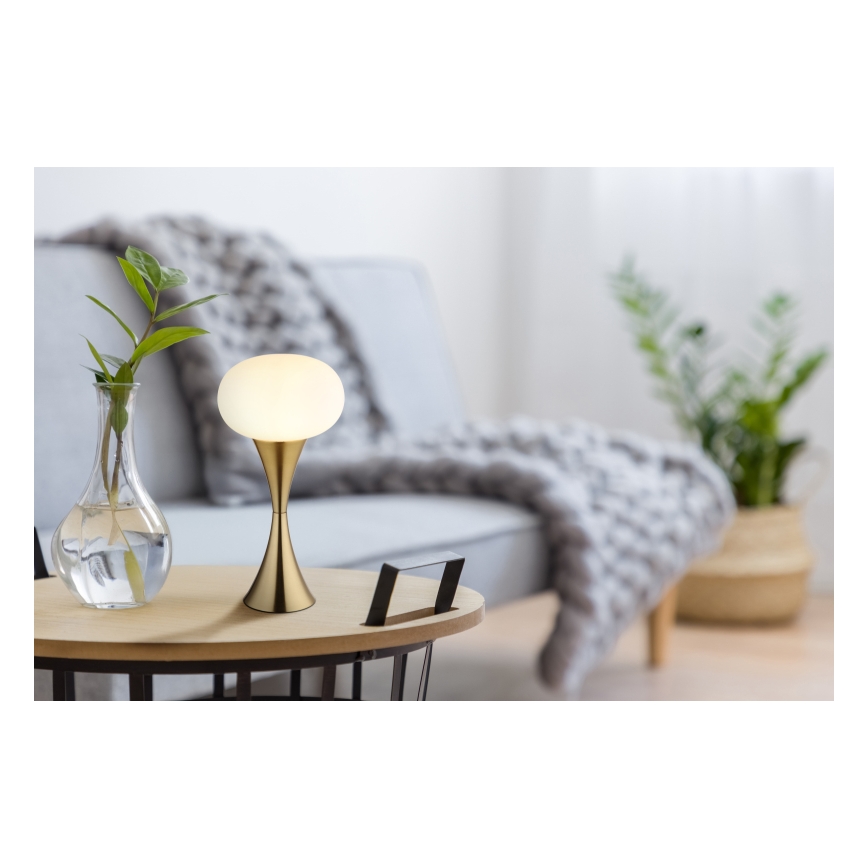 Stolná lampa s mliečnobielym tienidlom v tvare gule a zlatým podstavcom svieti na drevenom odkladacím stolíku. Lampa sa nachádza v obývacej izbe s pohovkou a rastlinami.