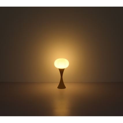 Stolová lampa s guľovou tienidlom svieti. Tienidlo je svetlé, stojan je hnedý a úzky. Lampa stojí na tmavej podlahe pred svetlou stenou.