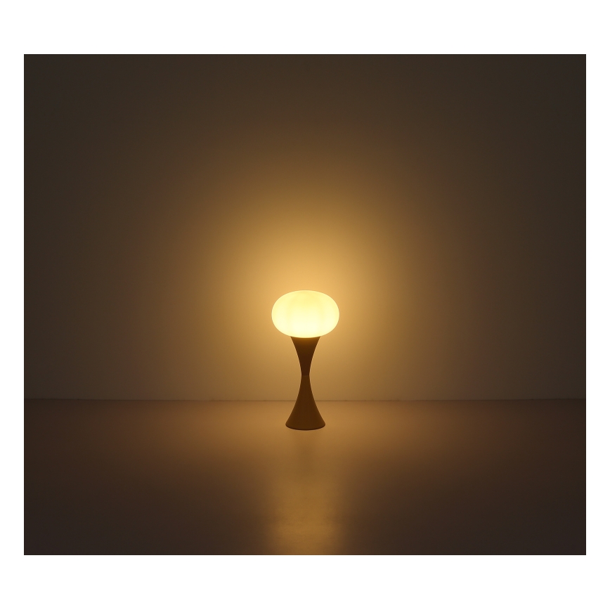 Stolová lampa s guľovou tienidlom svieti. Tienidlo je svetlé, stojan je hnedý a úzky. Lampa stojí na tmavej podlahe pred svetlou stenou.