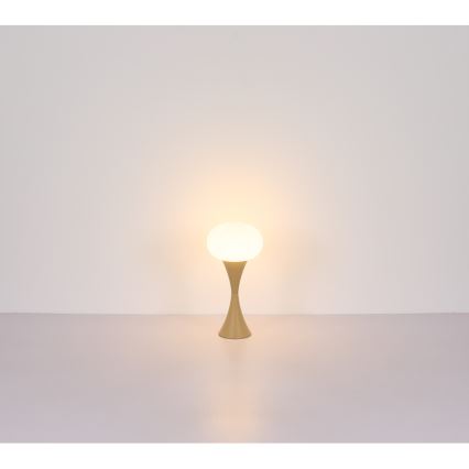 Stolná lampa s guľovou tienidlom svieti. Tienidlo je biele, stojan béžový. Lampa stojí na svetlej ploche pred svetlou stenou.