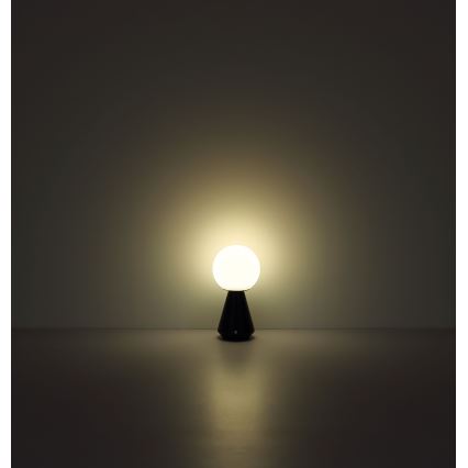 Stolová lampa svieti. Guľatá biela tienidlo je umiestnená na tmavom kužeľovitom podstavci. Lampa stojí na tmavej podlahe pred svetlou stenou.