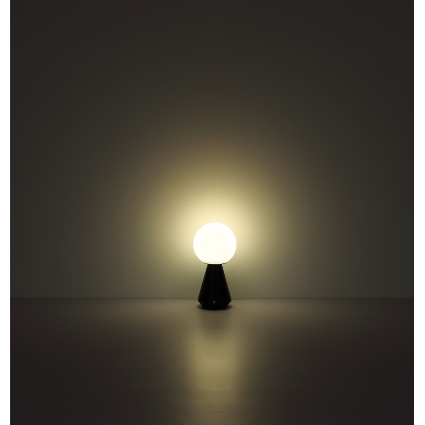 Stolová lampa svieti. Guľatá biela tienidlo je umiestnená na tmavom kužeľovitom podstavci. Lampa stojí na tmavej podlahe pred svetlou stenou.