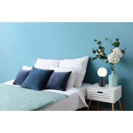 Stolová lampa s bielym guľovitým tienidlom a čiernou podstavou stojí na nočnom stolíku. Lampa je umiestnená v spálni pri posteli s bielou posteľnou bielizňou a tmavomodrými vankúšmi. Na nočnom stolíku je aj váza s kvetmi.