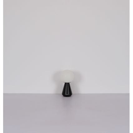 Stolná lampa s bielou guľovou tienidlom a čiernym kužeľovitým podstavcom stojí na svetlej ploche pred bielou stenou.