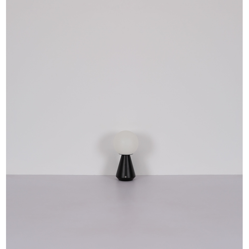 Stolná lampa s bielou guľovou tienidlom a čiernym kužeľovitým podstavcom stojí na svetlej ploche pred bielou stenou.