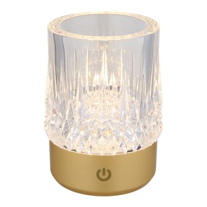 Stolová lampa svieti. Je vyrobená z priehľadného, fasetovaného skla na zlatom podstavci s vypínačom.