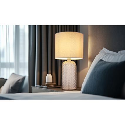 Stolová lampa s textilným tienidlom svieti na nočnom stolíku v spálni. Lampa má sklenenú podstavu s reliéfnym vzorom. Vedľa lampy je malá biela nádoba.