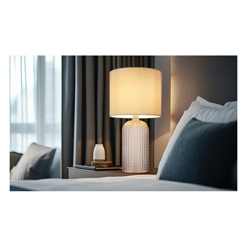 Stolová lampa s textilným tienidlom svieti na nočnom stolíku v spálni. Lampa má sklenenú podstavu s reliéfnym vzorom. Vedľa lampy je malá biela nádoba.