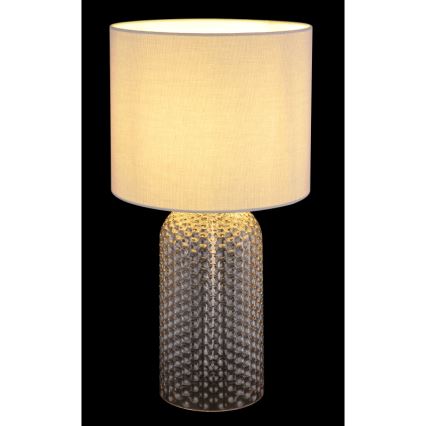 Stolová lampa so svetlo béžovým textilným tienidlom a priehľadnou sklenenou podstavou s reliéfnym vzorom guličiek, svieti. Je umiestnená na čiernom pozadí.