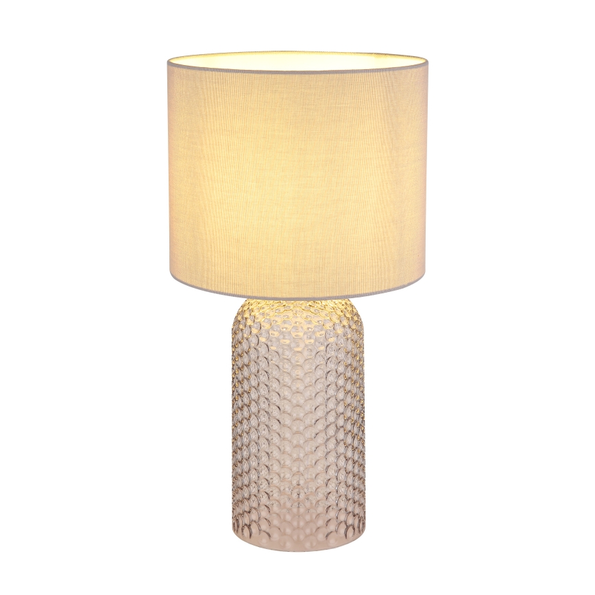 Stolová lampa s textilným tienidlom svetložltej farby svieti. Priesvitná sklenená základňa lampy je zdobená prelismi v tvare malých guličiek. Lampa je umiestnená na bielom pozadí.