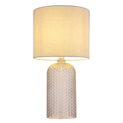 Stolová lampa s textilným tienidlom svetložltej farby stojí na podstavci z priehľadného skla s reliéfnym vzorom. Lampa je osvetlená.