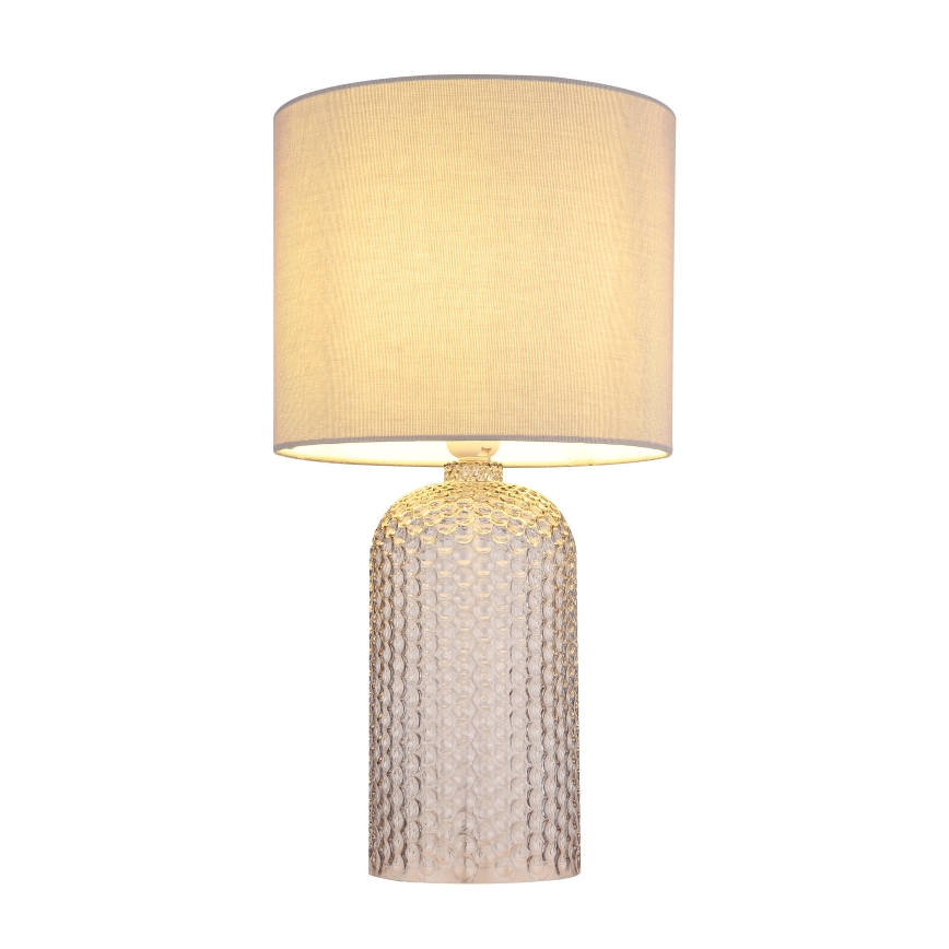 Stolová lampa s textilným tienidlom svetložltej farby stojí na podstavci z priehľadného skla s reliéfnym vzorom. Lampa je osvetlená.