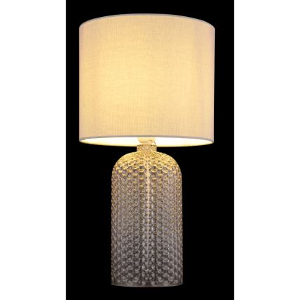 Stolová lampa svieti. Jej podstavec je z priehľadného skla s bodkovaným vzorom, tienidlo je svetlo béžové a látkové. Lampa je na čiernom pozadí.