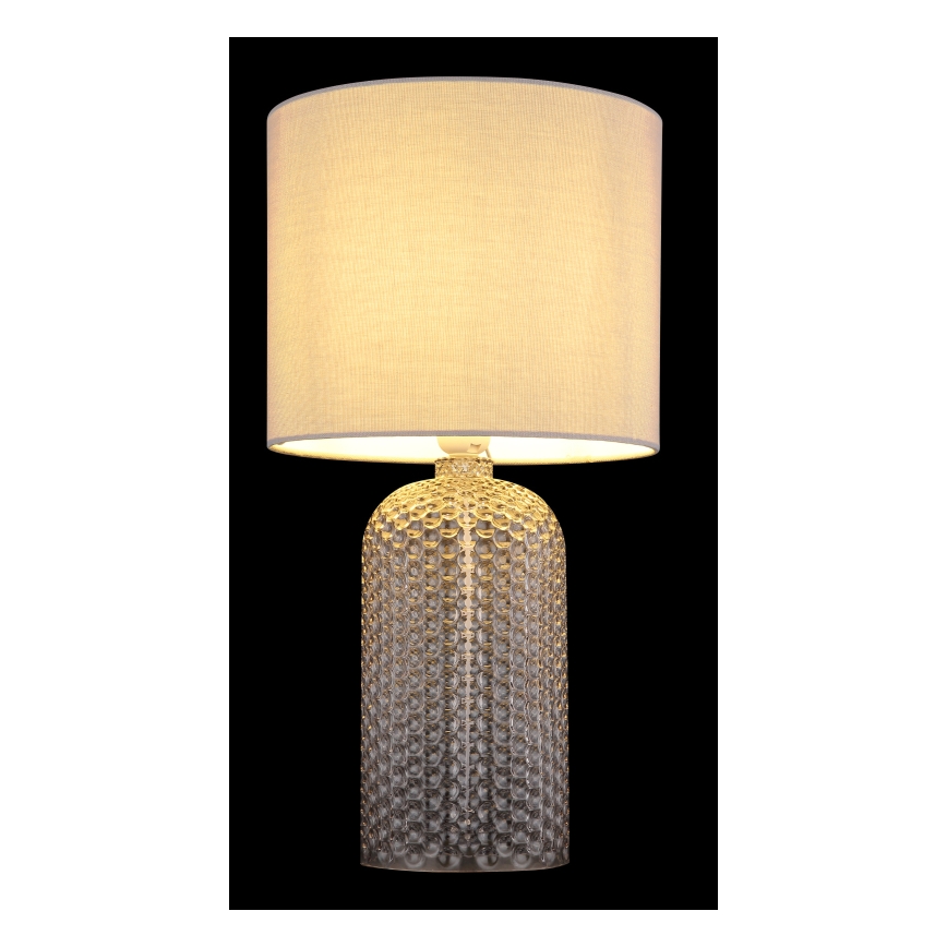 Stolová lampa svieti. Jej podstavec je z priehľadného skla s bodkovaným vzorom, tienidlo je svetlo béžové a látkové. Lampa je na čiernom pozadí.