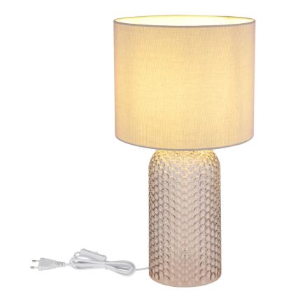Stolová lampa s textilným tienidlom svetložltej farby stojí na podstavci z priesvitného skla s reliéfnym vzorom. Je pripojená k elektrickej sieti pomocou vidlice.