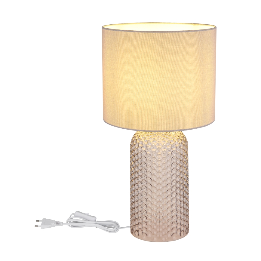 Stolová lampa s textilným tienidlom svetložltej farby stojí na podstavci z priesvitného skla s reliéfnym vzorom. Je pripojená k elektrickej sieti pomocou vidlice.