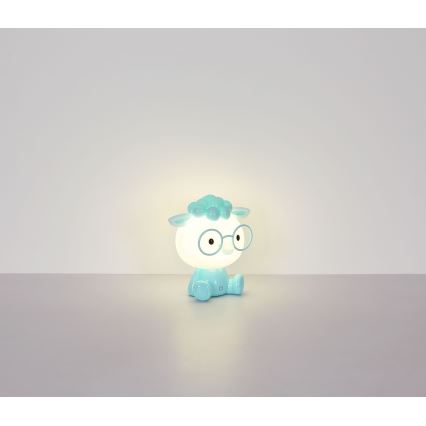 Svetlomodrá nočná lampa v tvare ovce s okuliarmi svieti. Sedí na svetlej podlahe pred svetlou stenou.