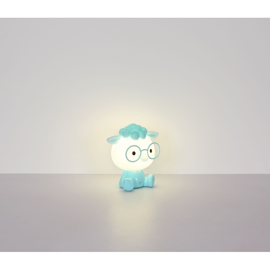 Svetlomodrá nočná lampa v tvare ovce s okuliarmi svieti. Sedí na svetlej podlahe pred svetlou stenou.
