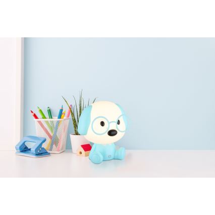 Svietiaca lampa v tvare psa s okuliarmi sedí na stole. Na stole sú tiež ceruzky, dierovač a kvetináč. Svetlomodrá lampa je umiestnená na bielom stole pred svetlomodrou stenou.