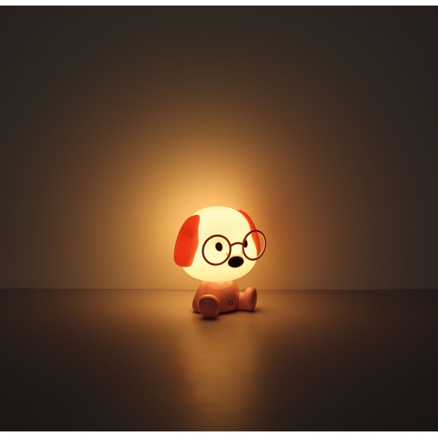 Svietiaca lampa v tvare psa s okuliarmi sedí na podlahe. Svieti mäkkým svetlom v tmavej miestnosti.