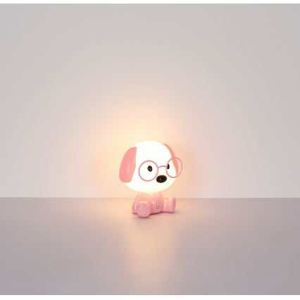 Ružová stolná lampa v tvare psa s okuliarmi svieti na svetlomodrej podlahe pred svetlou stenou.