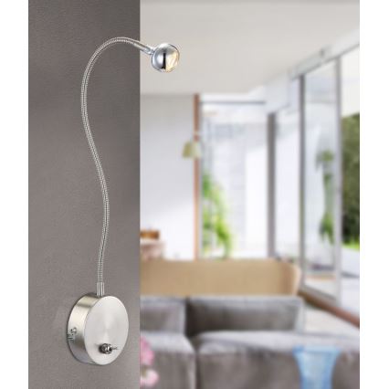 Nástenná lampa s ohybným ramenom svieti v interiéri. Je pripevnená na stenu a jej svetlo je smerované dopredu. V pozadí je rozmazané zobrazenie obývacej izby s pohovkou a oknami.