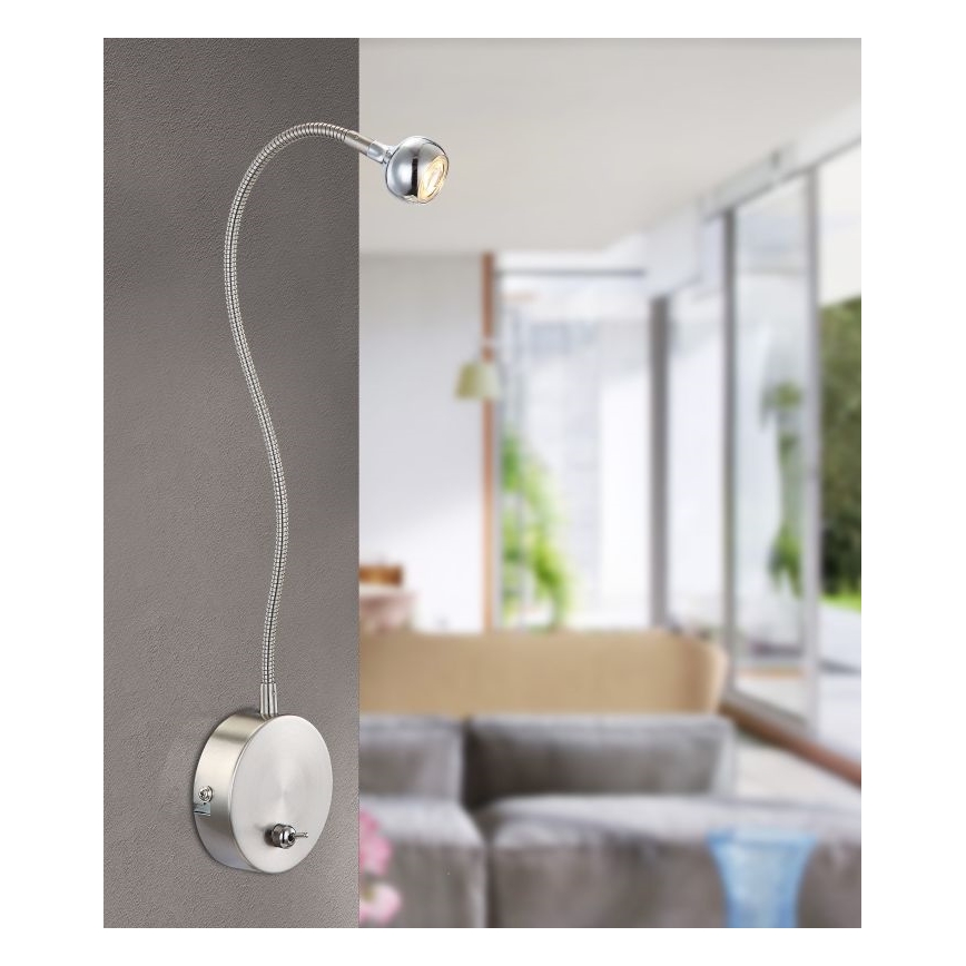 Nástenná lampa s ohybným ramenom svieti v interiéri. Je pripevnená na stenu a jej svetlo je smerované dopredu. V pozadí je rozmazané zobrazenie obývacej izby s pohovkou a oknami.