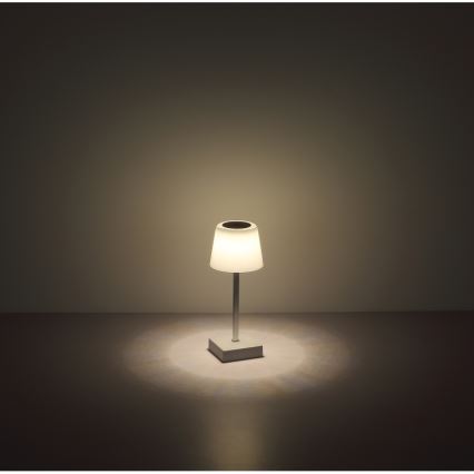 Malá stolová lampa s bielym tienidlom svieti v tmavej miestnosti. Je umiestnená na tmavej podlahe. Svetlo z lampy vytvára na podlahe svetelný kruh.