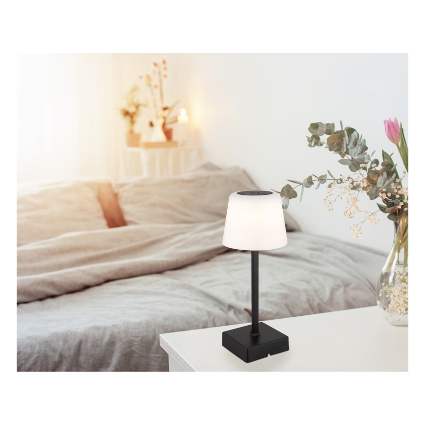 Malá stolová lampa s bielym tienidlom a čiernym stojanom svieti na nočnom stolíku v spálni. Lampa stojí na malej bielej nočnej stolíku pri posteli. V pozadí je rozostrená posteľ a váza s kvetmi.