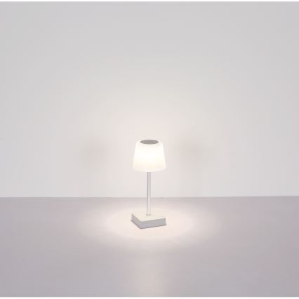 Malá stolová lampa s bielym tienidlom svieti na svetlom podklade pred bielou stenou. Lampa má tenký stĺpik a malú štvorcovú základňu.
