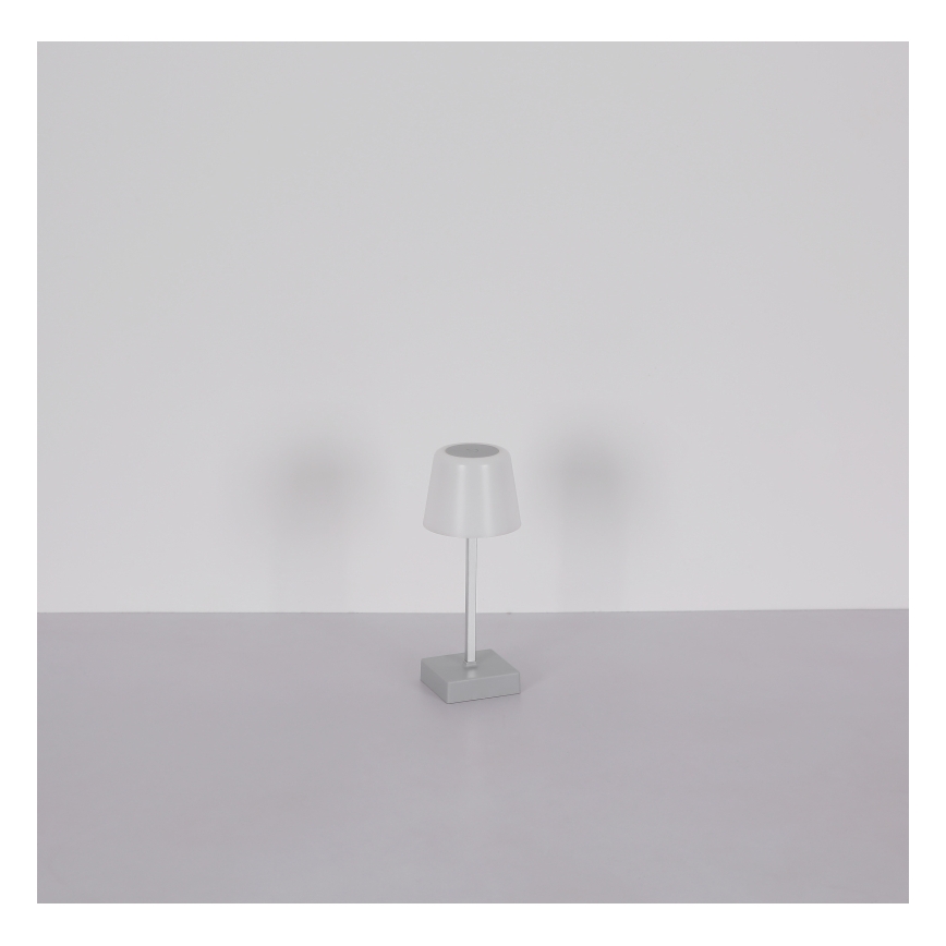 Malá stolová lampa s bielym tienidlom a sivým podstavcom stojí na svetlosivej ploche pred bielou stenou.