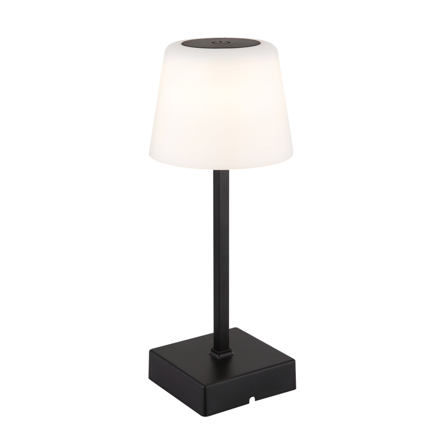 Stolová lampa svieti. Má biely tienidlo v tvare zrezaného kužeľa na čiernej kovovej stonke a štvorcovej základni.