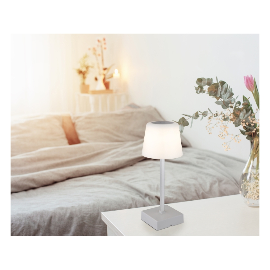 Stolová lampa s mäkkým svetlom svieti na nočnom stolíku v spálni. Lampa je sivá, má štíhle telo a tienidlo v tvare zrezaného kužeľa. V pozadí je posteľ s posteľnou bielizňou a kvetinová váza.