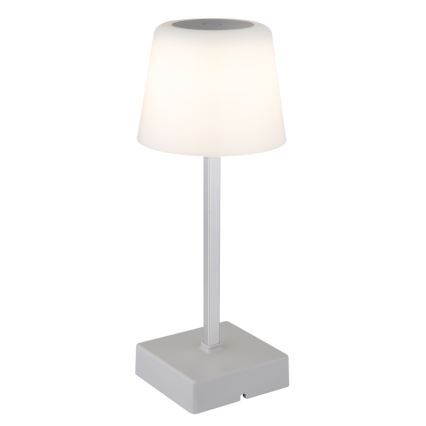 Stolná lampa svieti. Má sivý kovový stĺpik a biely tienidlo v tvare zrezaného kužeľa, ktoré je umiestnené na štvorcovej sivej základni.