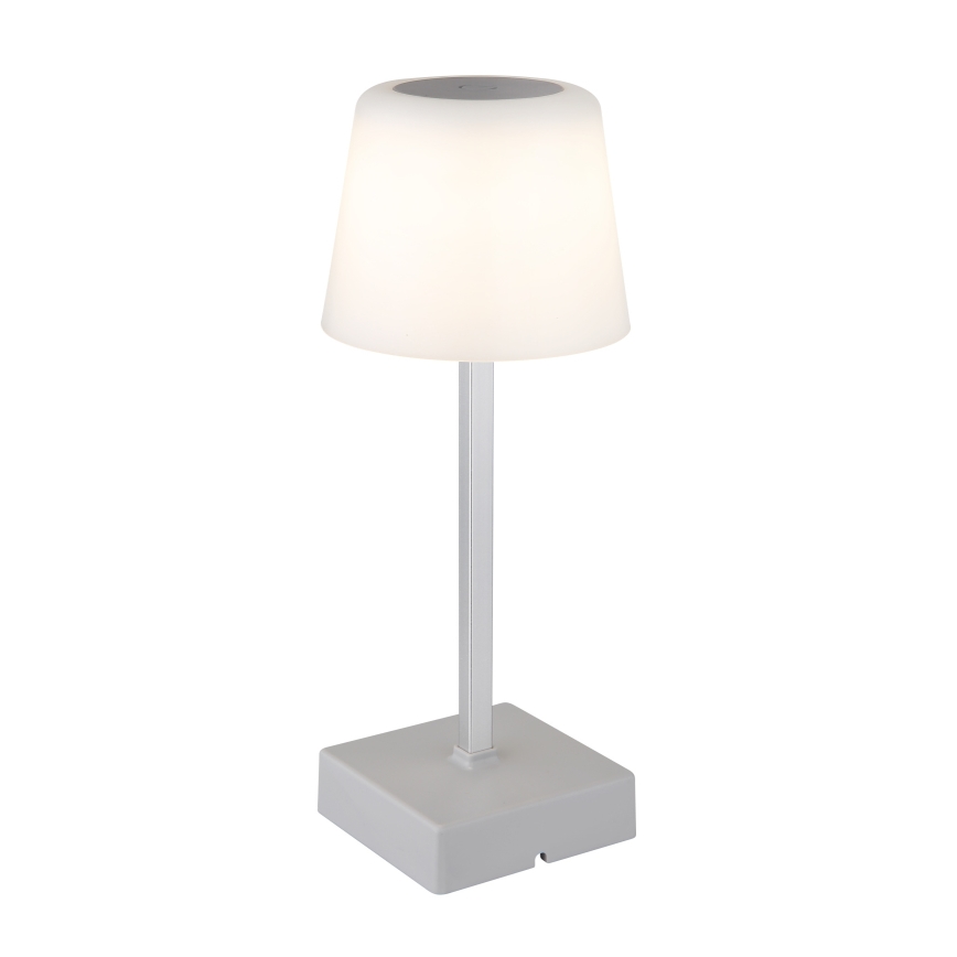 Stolná lampa svieti. Má sivý kovový stĺpik a biely tienidlo v tvare zrezaného kužeľa, ktoré je umiestnené na štvorcovej sivej základni.