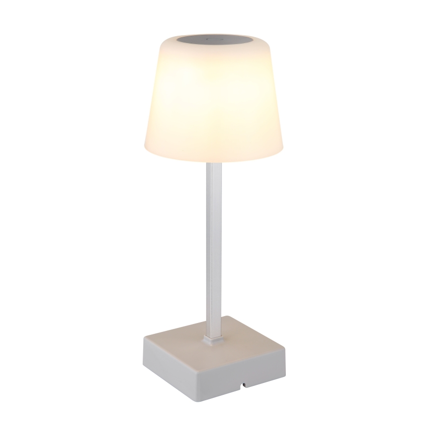Stolová lampa svieti. Má biely tienidlo tvaru zrezaného kužeľa a striebornú nohu na štvorcovej základni. Je umiestnená na bielom pozadí.