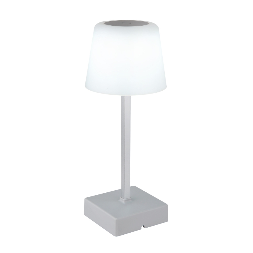 Stolová lampa s bielym tienidlom v tvare zrezaného kužeľa a striebornou nohou stojí na štvorcovej podstave. Svieti. Na vrchu tienidla je nenápadný tmavý kruh.