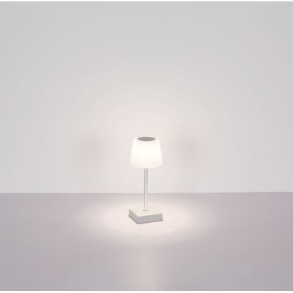 Malá stolová lampa s bielym tienidlom a kovovou nohou svieti na svetlej podlahe pred bielou stenou.