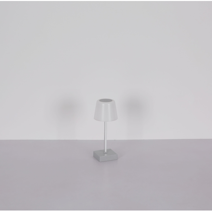 Malá stolová lampa s bielym tienidlom a sivou podstavou stojí na svetlosivej ploche pred bielou stenou. Lampa je umiestnená v strede obrazu.