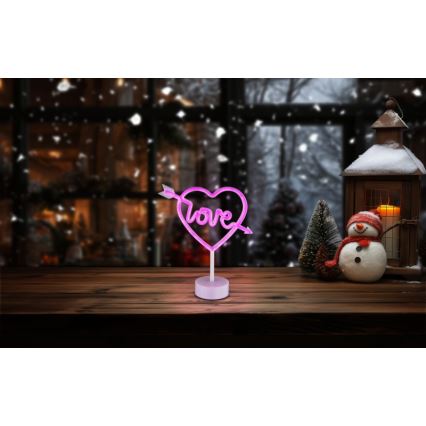 Neonové srdce s nápisom „Love“ a šípom svieti na drevenom stole pred zasneženým oknom a snehuliakom.