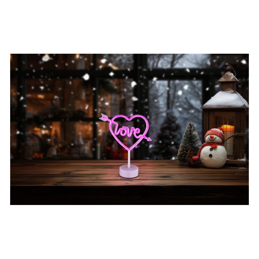 Neonové srdce s nápisom „Love“ a šípom svieti na drevenom stole pred zasneženým oknom a snehuliakom.