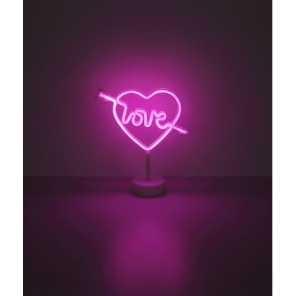 Neonové srdce s nápisom „love“ prebodnuté šípom svieti na bielom stojane. Svieti ružovým svetlom na tmavom podklade.